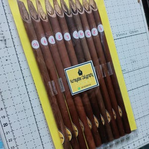 May include: A set of ten brown wooden calligraphy pens with pointed tips. The pens are numbered 1 through 10 and are packaged in a clear plastic bag on a yellow background. The bag has a label that reads "Nurhayatee Calligraphy Palani 0616209439".