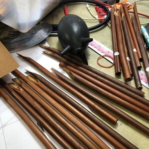 May include: A close-up of a group of brown wooden calligraphy pens with pointed tips. The pens are arranged in a fan shape on a white surface.