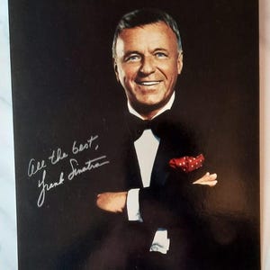 Peut inclure: Portrait de Frank Sinatra en smoking, souriant, avec une pochette rouge. L'image comprend sa signature et l'inscription "All the best". Le fond est noir.