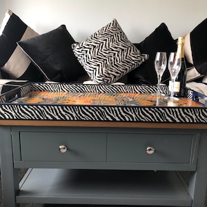 May include: A grey coffee table with two drawers and a lower shelf. A decorative tray with a zebra print border and colourful centre sits on top. Champagne glasses and a bottle of champagne are on the tray. Black and white cushions are on the sofa behind the table.