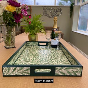 May include: A rectangular serving tray with dark green edges and a light green leaf pattern on the base and sides. The tray features two cutout handles and measures 60cm x 40cm. The interior surface displays a repeating botanical print of green leaves against a light background, providing a flat area for carrying items.