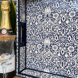 May include: A rectangular serving tray featuring a dark navy blue and cream floral pattern with intricate botanical motifs. The tray has high sides, integrated cutout handles, and a glossy finish. A champagne bottle and glass are positioned nearby.