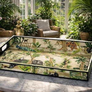 May include: Rectangular serving tray with a jungle-themed design. The tray has a black border and handles, with a cream interior showcasing peacock, tiger, and monkey illustrations. The tray is placed on a marble table.