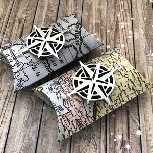 May include: Two pillow-shaped gift boxes with a vintage map design. Each box is topped with a white compass rose. The boxes are on a wooden surface.