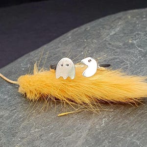 May include: A pair of silver earrings featuring a ghost and a Pac-Man design. The earrings are on a yellow feather.