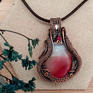 Puede incluir: Collar con colgante artesanal con una piedra en forma de lágrima en tonos rojos y blancos, envuelta en alambre de cobre. El colgante cuelga de un cordón de cuero marrón, mostrando un diseño artístico único. Un pequeño cristal y una cuenta roja acentúan el trabajo de alambre.