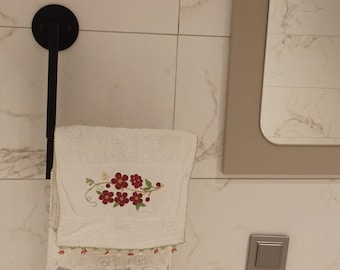 May include: A white towel with a floral embroidery design hangs on a black towel rack in a bathroom. The towel has a crocheted border and is folded in half. The bathroom has white marble tiles and a white sink.