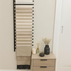 May include: A black metal scarf rack displays a collection of neutral-toned scarves. The scarves range in colour from cream to beige, with the bottom scarf featuring a fringe. A wooden bedside table with three drawers and a vase with dried flowers are also visible.