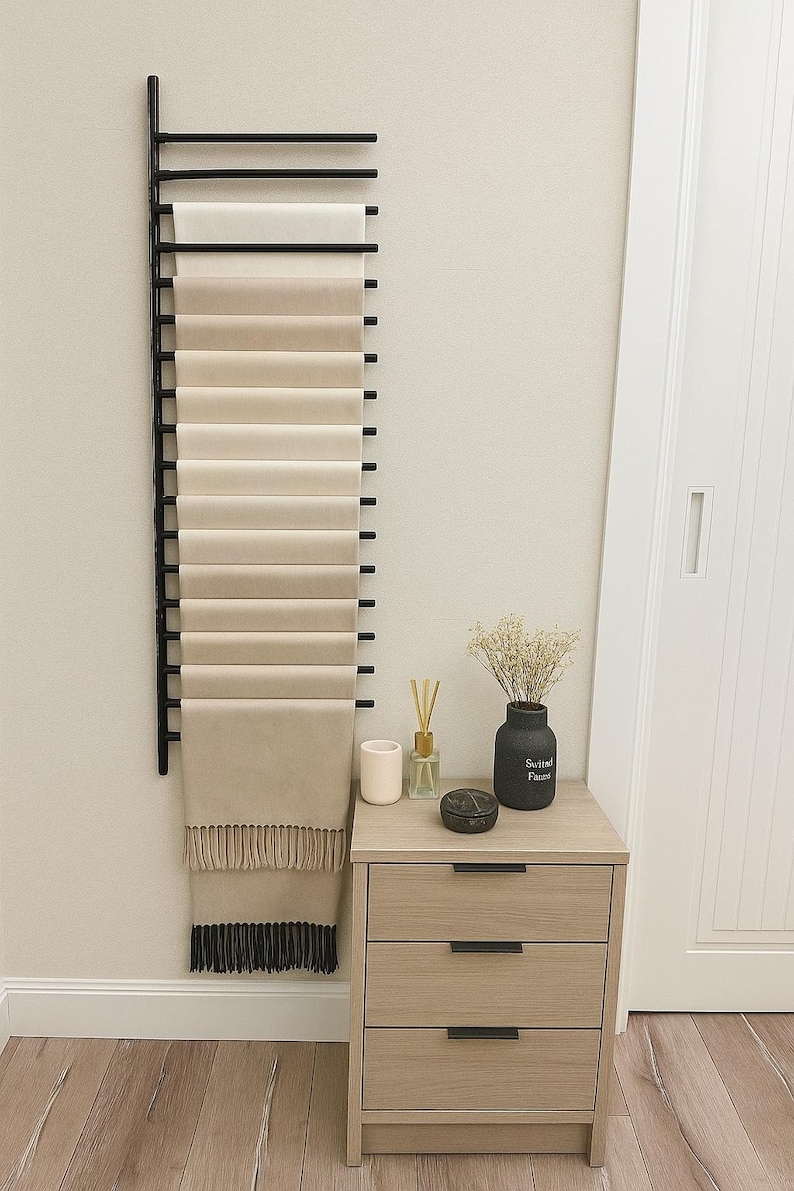 May include: A black metal scarf rack displays a collection of neutral-toned scarves. The scarves range in colour from cream to beige, with the bottom scarf featuring a fringe. A wooden bedside table with three drawers and a vase with dried flowers are also visible.