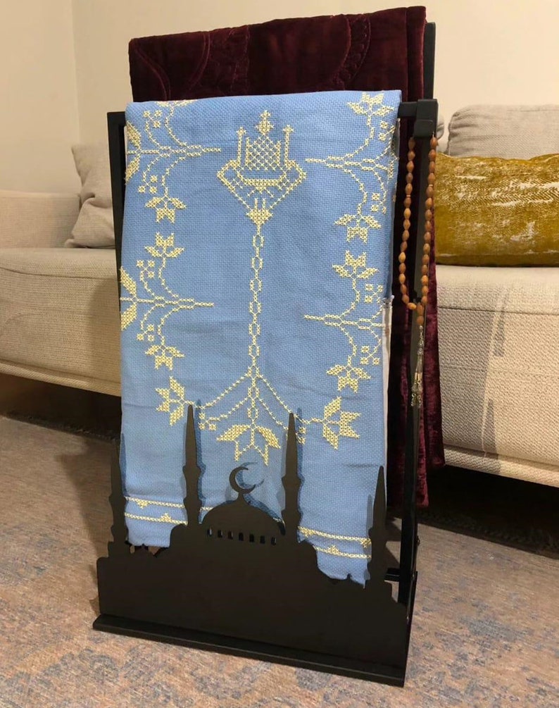 May include: A black metal prayer rug stand with a blue and gold prayer rug hanging on it. The stand features a silhouette of a mosque with a crescent moon.