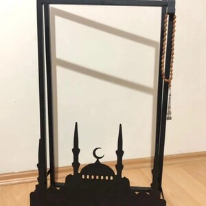 May include: Black metal stand with a silhouette of a mosque and a crescent moon. A brown wooden prayer bead necklace hangs on the side.