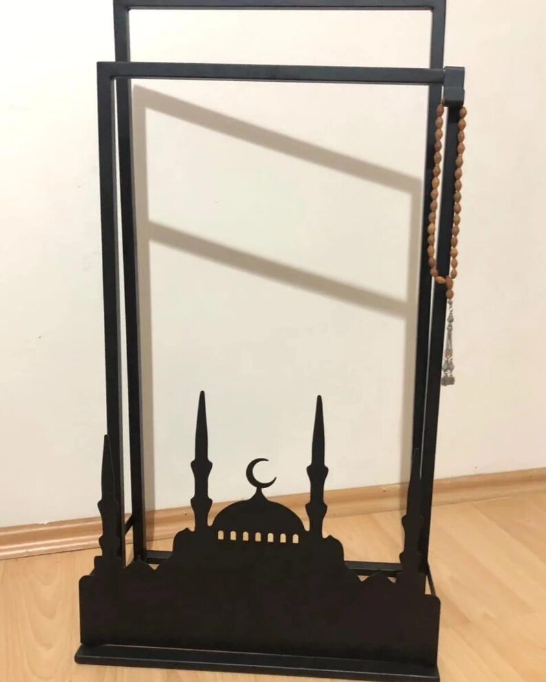 May include: Black metal stand with a silhouette of a mosque and a crescent moon. A brown wooden prayer bead necklace hangs on the side.