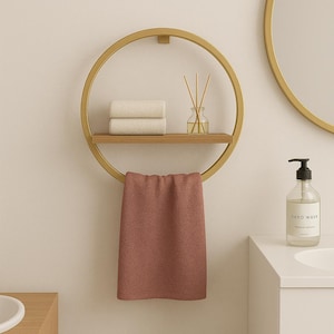 May include: A gold-colored, circular towel rack with a wooden shelf holding two rolled white towels and a glass diffuser. A dusty rose towel hangs from the bottom. A round mirror and a hand wash bottle are visible.