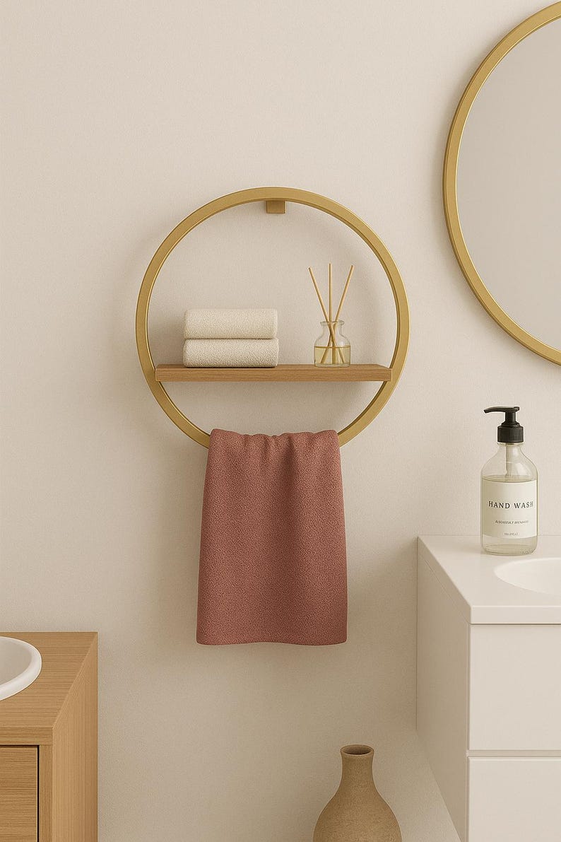 May include: A gold-colored, circular towel rack with a wooden shelf holding two rolled white towels and a glass diffuser. A dusty rose towel hangs from the bottom. A round mirror and a hand wash bottle are visible.