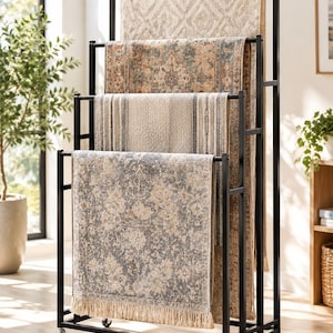 May include: A black metal tiered display rack on wheels, holding four rectangular rugs with various patterns and textures. The rugs feature neutral tones, including beige, cream, blue, and gray, with one rug displaying a fringed bottom edge.