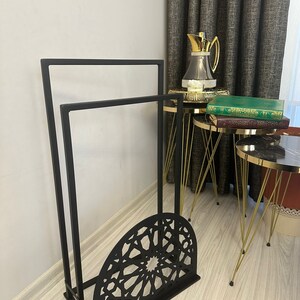 May include: Black metal stand with two shelves and a decorative, geometric, cut-out design. The stand is on a wooden floor.