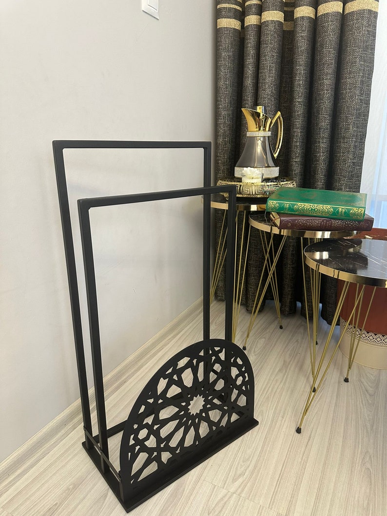 May include: Black metal stand with two shelves and a decorative, geometric, cut-out design. The stand is on a wooden floor.