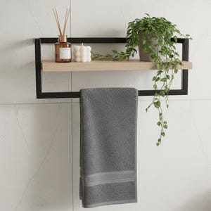 May include: A modern bathroom shelf with a black metal frame and a light wood shelf. The shelf holds a potted plant, a candle, and a diffuser. A gray towel hangs below. The shelf is mounted on a white tiled wall.