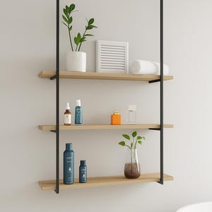 Może przedstawiać: Trzypoziomowa, wisząca półka łazienkowa z czarnymi metalowymi wspornikami i jasnymi drewnianymi półkami. Półki zawierają różne kosmetyki, rośliny w białych i szklanych wazonach oraz zwinięty biały ręcznik. Nowoczesny i minimalistyczny design.