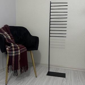 May include: A black metal stand with multiple horizontal bars for hanging items. The stand has a rectangular base and is standing on a light wood floor.