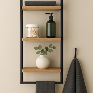 May include: A contemporary bathroom storage unit with a black metal frame and wooden shelves. It features a grey towel, a glass jar of cotton buds, a green soap dispenser, a white vase with foliage, and a hanging grey towel.