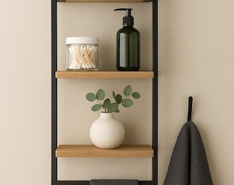 May include: A modern bathroom shelf unit with a black metal frame and wooden shelves. It holds a gray towel, a glass jar of cotton swabs, a green soap dispenser, a white vase with greenery, and a hanging gray towel.