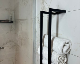 May include: Black metal towel rack with three white towels rolled up and placed on the rack. The rack is mounted on a white tiled wall.