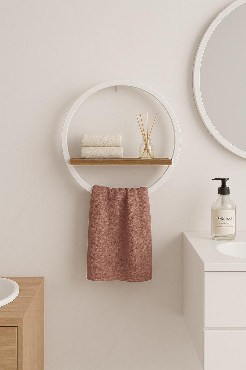 May include: A modern bathroom shelf with a white circular frame, a wooden shelf, and a hanging towel. The shelf holds two folded white towels and a glass diffuser. A pink towel hangs below. A bottle of hand wash is on the counter.