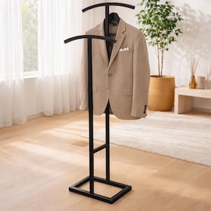 Könnte beinhalten: Ein schwarzer Herrendiener aus Metall mit rechteckigem Fuß, vertikalem Ständer und zwei geschwungenen horizontalen Stangen zum Aufhängen von Kleidung. Ein beigefarbener Blazer mit weißem Einstecktuch hängt an der oberen Stange des Ständers.