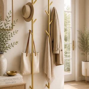 K&ouml;nnte beinhalten: Zwei goldfarbene Metall-Kleiderst&auml;nder an einer Wand, jeder mit mehreren Zweigen zum Aufh&auml;ngen von Gegenst&auml;nden. Ein beiger Hut, eine Tragetasche, ein Schal und ein Mantel sind ausgestellt. Ein kleiner Beistelltisch mit B&uuml;chern und einer Vase ist ebenfalls zu sehen.