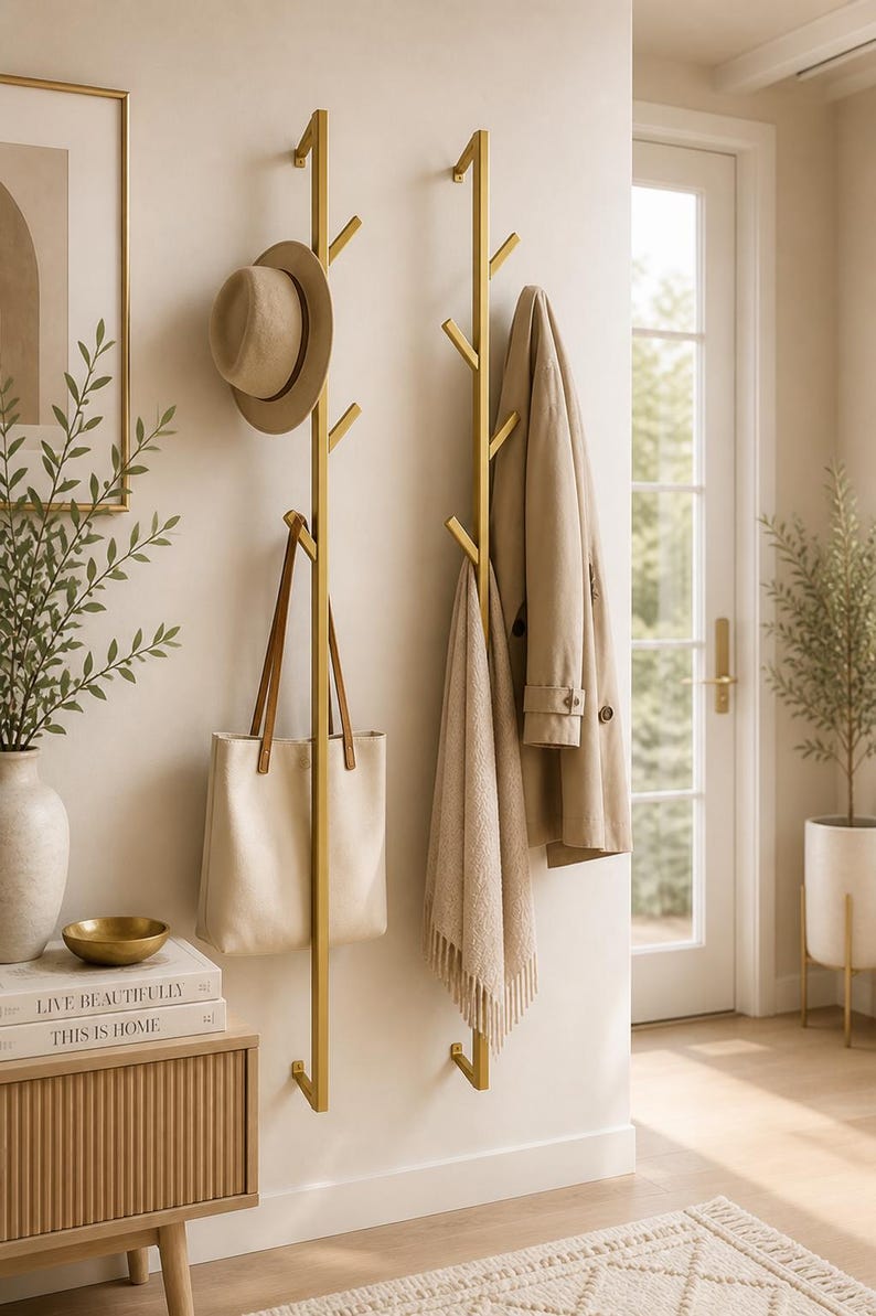 K&ouml;nnte beinhalten: Zwei goldfarbene Metall-Kleiderst&auml;nder an einer Wand, jeder mit mehreren Zweigen zum Aufh&auml;ngen von Gegenst&auml;nden. Ein beiger Hut, eine Tragetasche, ein Schal und ein Mantel sind ausgestellt. Ein kleiner Beistelltisch mit B&uuml;chern und einer Vase ist ebenfalls zu sehen.