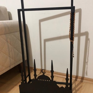 May include: Black metal stand with a mosque silhouette design. The stand has two horizontal bars and a small shelf. A brown wooden prayer bead necklace hangs from the top bar.