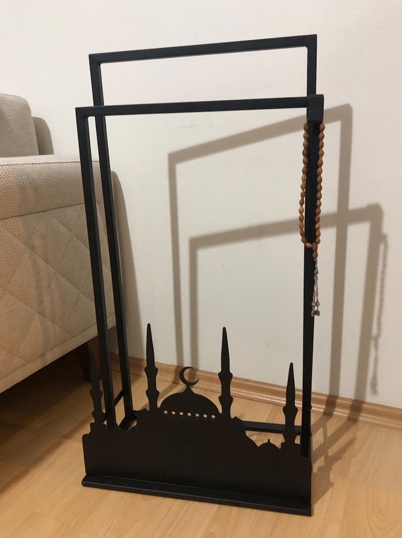 May include: Black metal stand with a mosque silhouette design. The stand has two horizontal bars and a small shelf. A brown wooden prayer bead necklace hangs from the top bar.