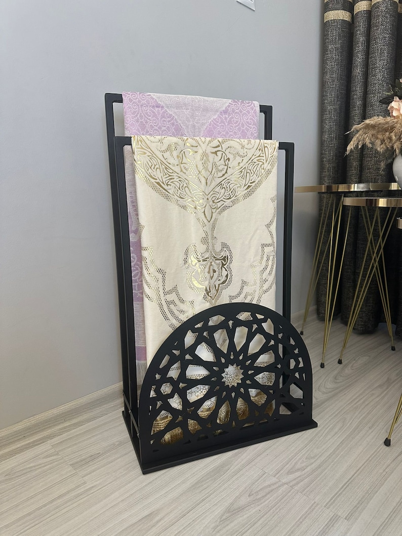 May include: Black metal prayer rug stand with an intricate geometric design. The stand has two shelves for storing prayer rugs.