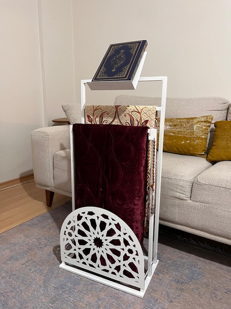 May include: White metal prayer stand with a burgundy prayer rug, a patterned rug, and a blue book on top. The stand has a decorative cut-out design in the shape of an arch.