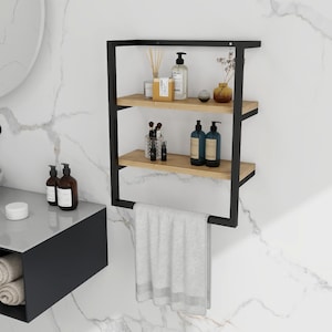 May include: A black metal and wood bathroom shelf with three levels. The top two shelves hold bottles, jars, and decorative items. A grey towel hangs from the bottom. The shelf is mounted on a white marble-patterned wall.