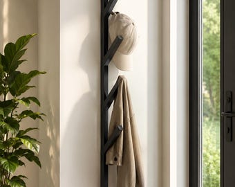 May include: A vertical black wall-mounted coat rack with five angled hooks. A beige baseball cap hangs from the third hook, and a tan long-sleeved shirt hangs from the fourth hook. The rack has a simple, linear design with a matte finish.