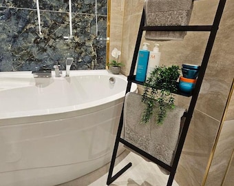 May include: A black metal leaning ladder shelf in a bathroom setting. The shelf holds folded gray towels, toiletries, and a small potted plant. The bathroom has a white bathtub and marble-look tile.