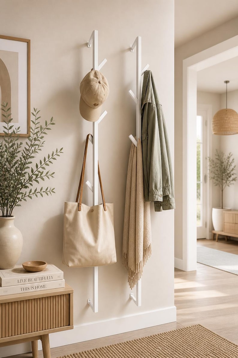 K&ouml;nnte beinhalten: Ein wei&szlig;er, an der Wand befestigter Kleiderst&auml;nder mit mehreren Zweigen, an dem eine beige Tragetasche, eine beige Baseballkappe, eine hellgr&uuml;ne Jacke und ein beiger Schal h&auml;ngen. Der St&auml;nder ist an einer hellbeigen Wand befestigt, mit einem h&ouml;lzernen Beistelltisch und einem gerahmten Bild im Hintergrund.