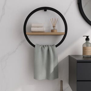 May include: A modern black metal towel rack with a circular design. It features a wooden shelf holding rolled white towels and a diffuser. A gray towel hangs from the bottom. The rack is mounted on a white marble wall, with a matching mirror and cabinet.