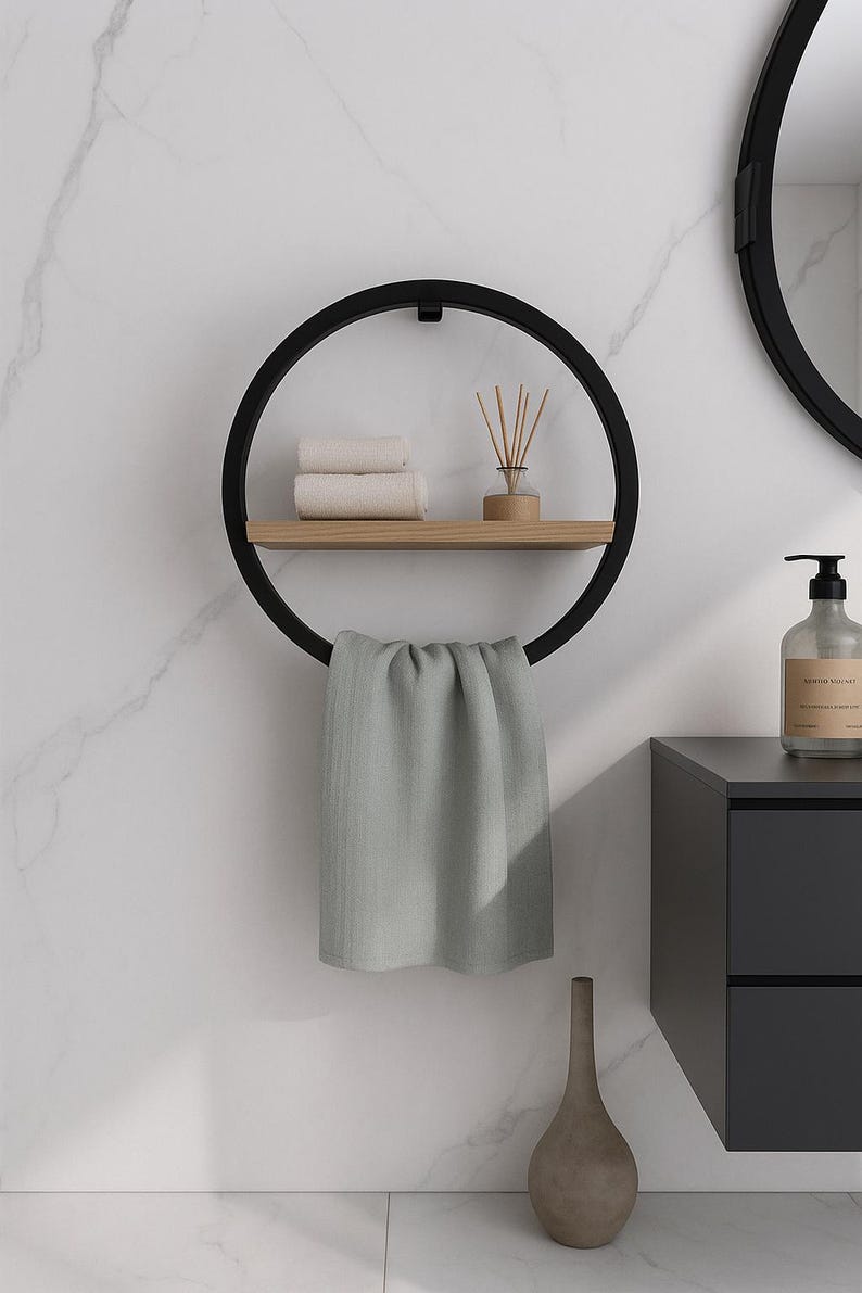 May include: A modern black metal towel rack with a circular design. It features a wooden shelf holding rolled white towels and a diffuser. A gray towel hangs from the bottom. The rack is mounted on a white marble wall, with a matching mirror and cabinet.