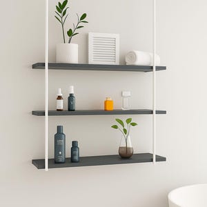 May include: A modern, three-tiered floating shelf unit with white support poles. The shelves hold various bathroom accessories, including plants, bottles, and a rolled towel. The shelves are dark gray.