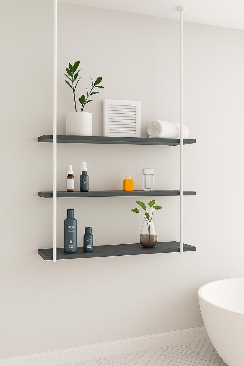 May include: A modern, three-tiered floating shelf unit with white support poles. The shelves hold various bathroom accessories, including plants, bottles, and a rolled towel. The shelves are dark gray.