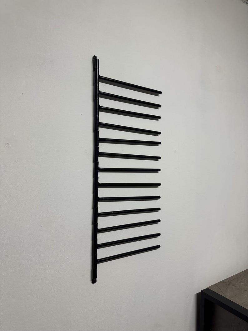 May include: A black metal wall-mounted rack with several horizontal bars. The rack is designed for storage and is fixed to a white wall. The design is minimalist and modern, suitable for various storage needs.