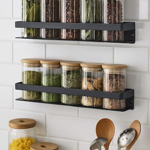 Peut inclure: Scène de cuisine avec deux étagères à épices en métal noir fixées à un mur carrelé blanc. Chaque étagère contient plusieurs bocaux en verre transparent avec des couvercles en bois, remplis de diverses épices. D'autres bocaux et ustensiles en bois sont également visibles.