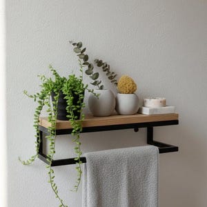 May include: A modern bathroom shelf with a black metal frame and a light wood shelf. It holds a gray towel, a potted plant, two small white vases, a yellow sponge, and two bars of soap. The shelf is mounted on a white wall.