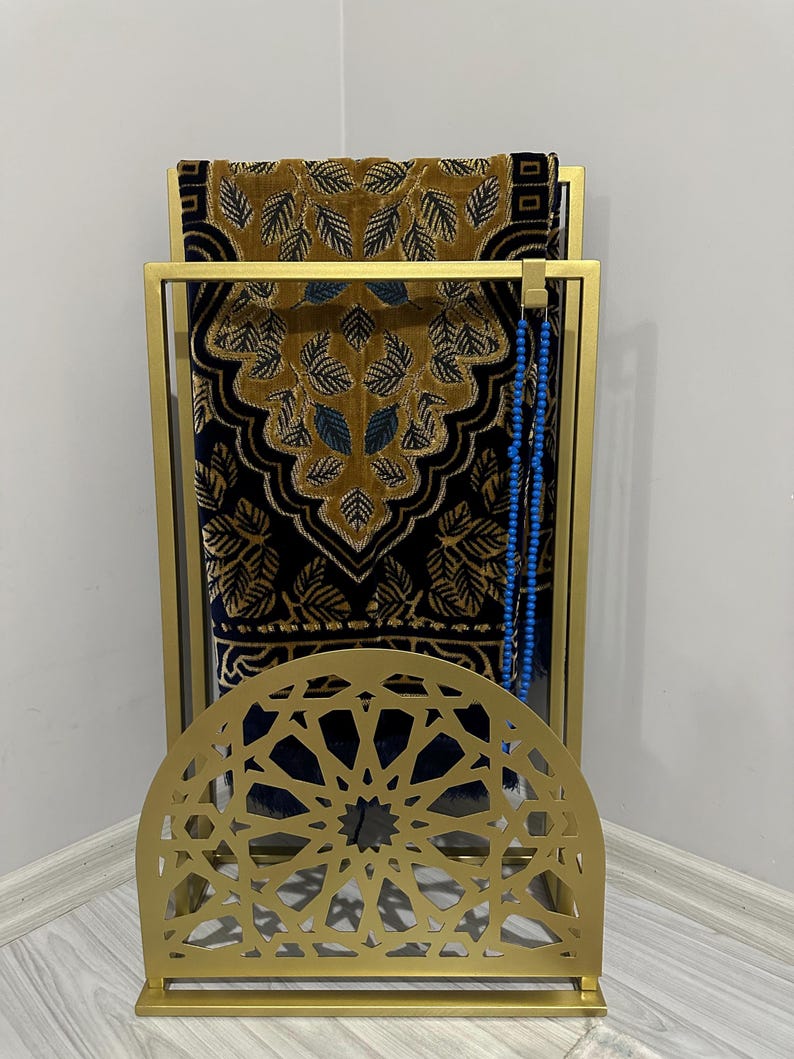 May include: A gold metal prayer rug stand with an intricate geometric design. The stand has a shelf for storing a prayer rug and a hook for hanging prayer beads. The rug is brown and black with a floral pattern.