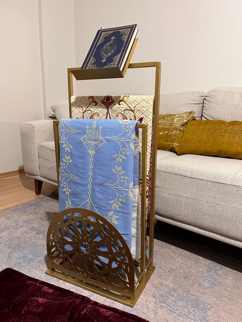 May include: A gold metal prayer stand with a blue prayer rug and a book on top. The stand has a decorative design and a shelf for the book.
