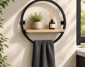 May include: A circular black metal wall shelf featuring a horizontal light wood plank. The shelf holds a small potted green plant and a glass bottle with reed diffusers. A dark gray waffle-textured towel hangs over the bottom of the metal frame.