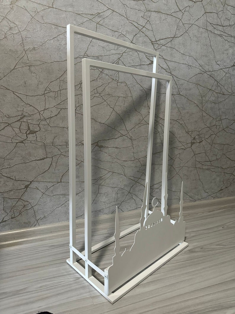 May include: White metal stand with three tiers, featuring a silhouette of a mosque with minarets. The stand is designed for displaying items or as a decorative piece.